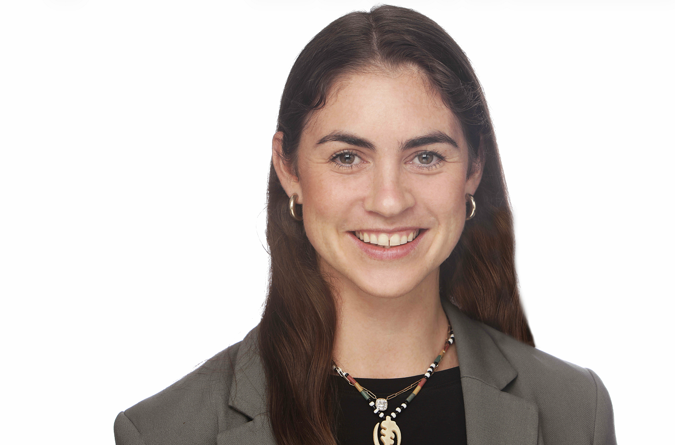 Image of Monica smiling to camera. They have long brown hair and are wearing a black shirt and gray blazer with a color necklace in front of a white background.